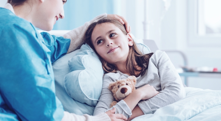 Barn i hospitalseng med bamse i favn og kvinde ved siden af