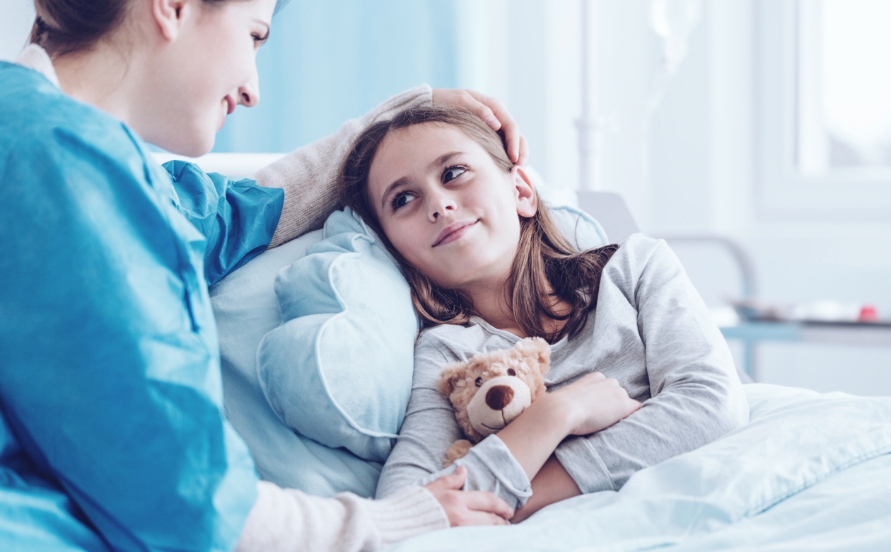 Barn i hospitalseng med bamse i favn og kvinde ved siden af