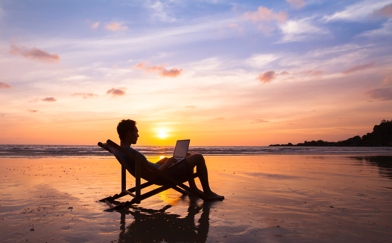 Person sidder i strandstol med laptop på stranden ved solnedgang