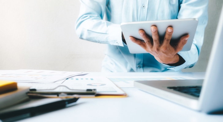 Person analyserer data på ipad med papirer og laptop på bord foran