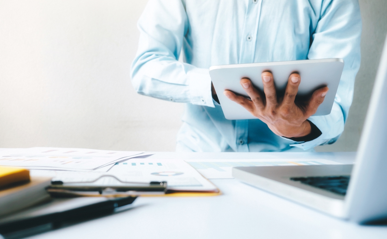 Person analyserer data på ipad med papirer og laptop på bord foran