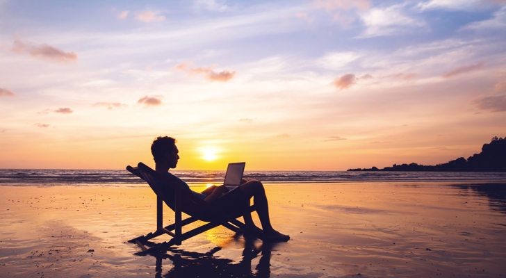 Mand med computer siddende i strandstol på stranden ved solnedgang
