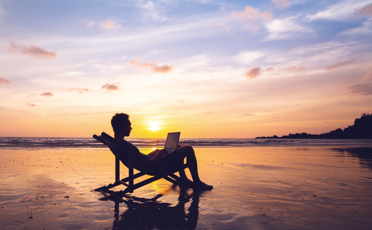 Mand med computer siddende i strandstol på stranden ved solnedgang