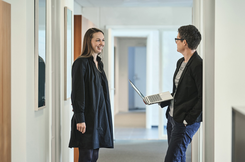 Ditte Marie Hansen og Jeanette Dich Sørensen med bærbar i hånden står og taler sammen på gang